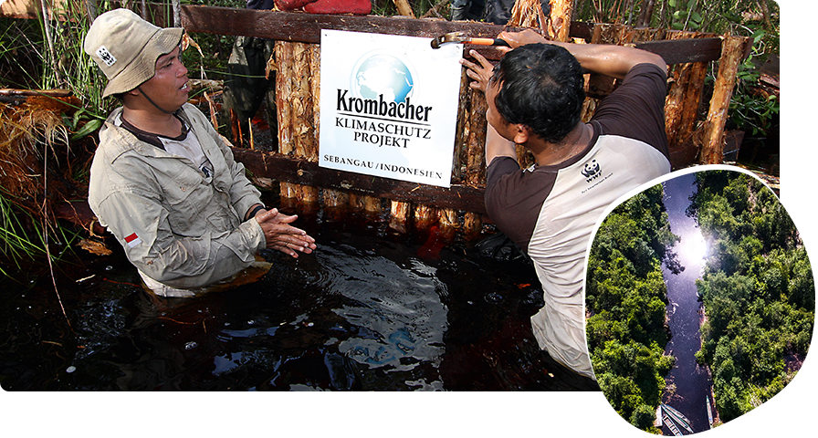 Zwei Personen stehen im Fluss und bringen ein Schild an einem Zaun an, mit der Aufschrift: Krombacher Klimaschutz Projekt.