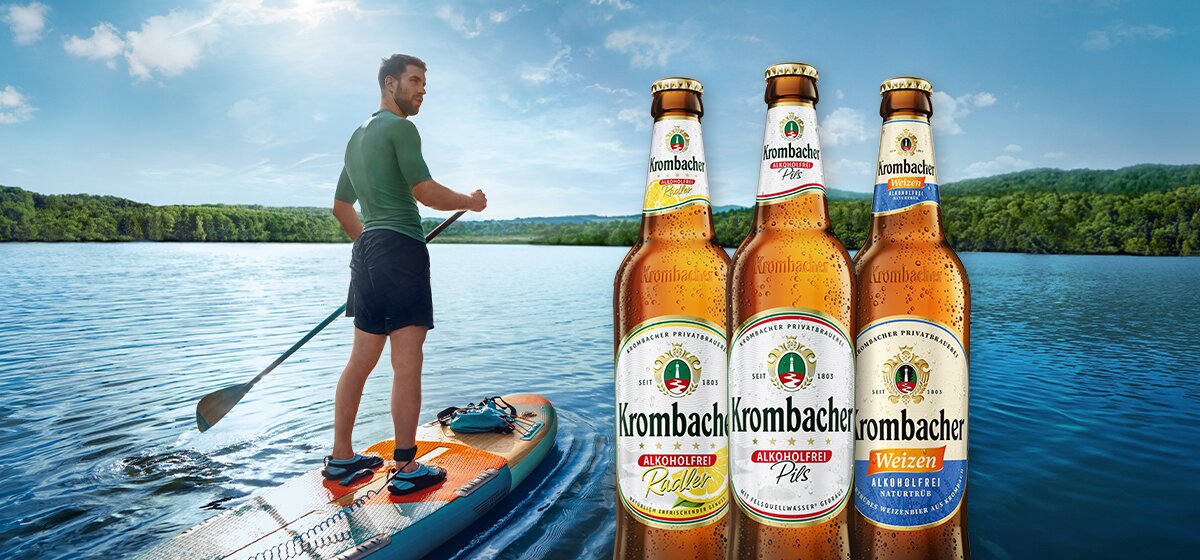 Mann steht auf einem Stand-Up-Paddle-Board auf einem See, drei Flaschen Krombacher Bier in den Sorten Radler, Pils und Weizen im Vordergrund.