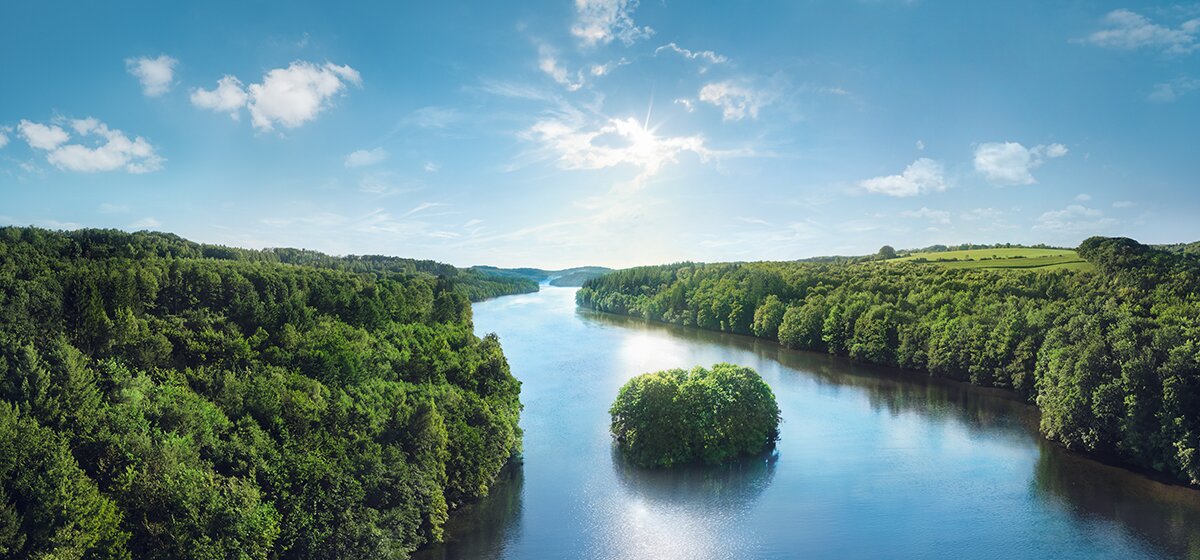 Abbildung des Krombacher Sees mit einer Insel, umgeben von dichtem Wald unter blauem Himmel mit einzelnen Wolken.