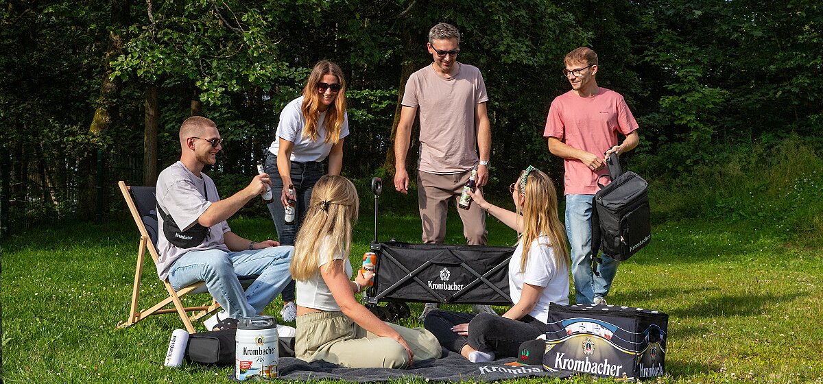 Gruppe von jungen Menschen sitzen in Freiem auf einer Wiese vor Bäumen und stoßen mit Krombacher an. Zu sehen sind weitere Krombacher Produkte.