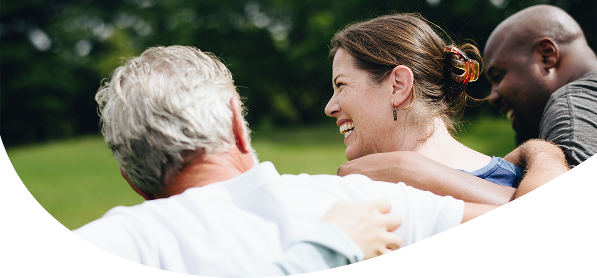 Zwei Männer und eine Frau legen sich gegenseitig die Arme über die Schultern und lächeln. Sie stehen im freien auf einer Wiese.