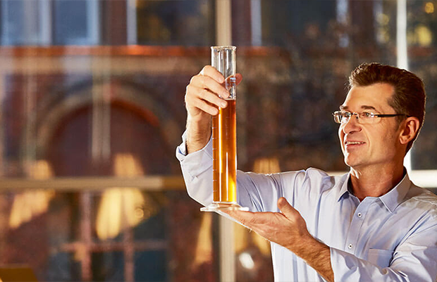 Zu sehen ist Manfred Schmidt der Geschäftsführer der Produktion und Technik. Er hält ein transparentes Messglas mit Pils darin ins Licht und begutachtet es.