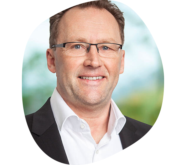 Portrait von Wolfgang Schötz, Leiter Nachhaltigkeitsmanagement. Er hat braune Haare, trägt eine Brille, einen weißen Hemdkragen, schwarzen Anzug und lächelt.