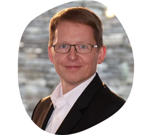 Mann in schwarzem Sakko und weißem Hemd, mit Brille vor unscharfem Hintergrund.