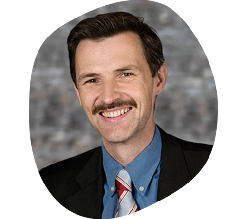 Mann mit Braunen Haaren und Schneuzer, trägt ein blaues Hemd mit Krawatte und schwarzem Sakko.