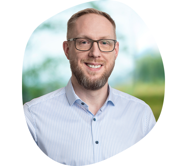 Portrait von Christian Zimmer, Leiter der Produktion. Er hat blonde Haare, trägt eine Brille und einen Dreitagebart. Er tägt ein hellblaues-weiß gestreiftes Hemd und lächelt in die Kamera.