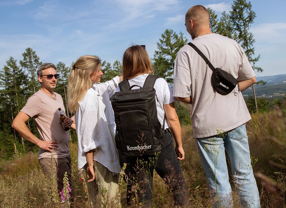 Zwei Männer und zwei Frauen stehen auf einer Wiese, umgeben von Bäumen und Hügeln. 