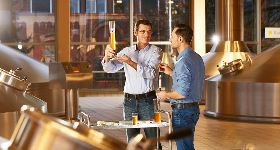 Zwei Männer in einer Brauerei halten ein Glas Bier und stehen vor großen Brautanks.