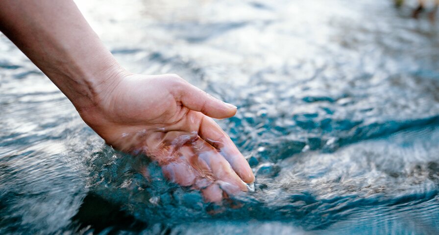 Hand schöpft klares Wasser aus einem fließenden Gewässer.