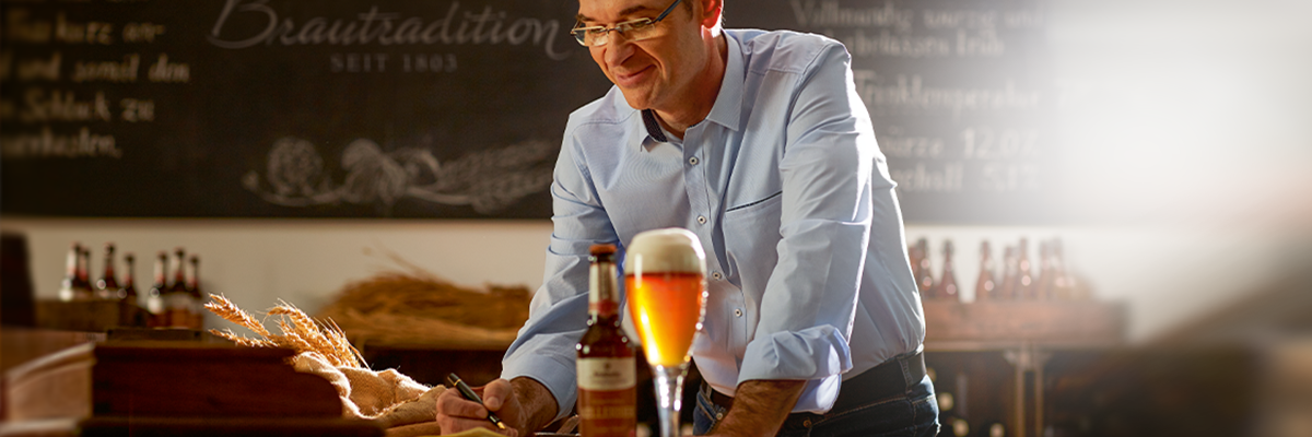 Ein Mann in einem hellblauen Hemd steht an einem Tisch, schreibt und hat ein Glas Krombacher Kellerbier sowie eine Flasche auf dem Tisch.