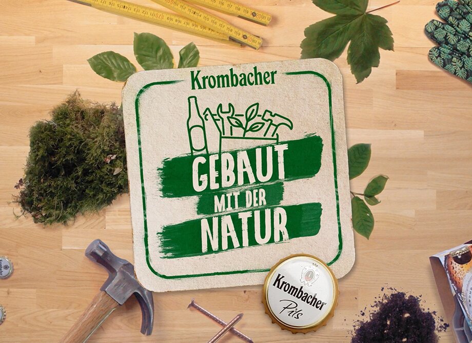 Das Bild zeigt ein Krombacher-Logo mit dem Text 'GEBAUT MIT DER NATUR' auf einem Untergrund aus Holz. Um das Logo sind verschiedene Gartenwerkzeuge, Pflanzen und eine Krombacher-Bierdose angeordnet.