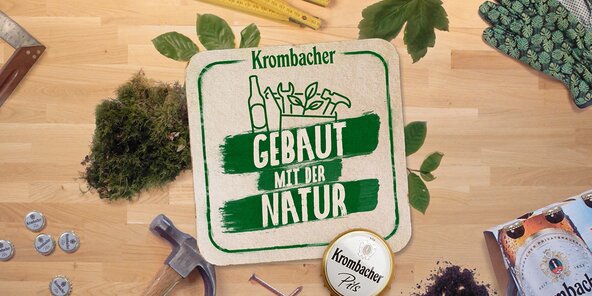 Das Bild zeigt ein Krombacher-Logo mit dem Text 'GEBAUT MIT DER NATUR' auf einem Untergrund aus Holz. Um das Logo sind verschiedene Gartenwerkzeuge, Pflanzen und eine Krombacher-Bierdose angeordnet.