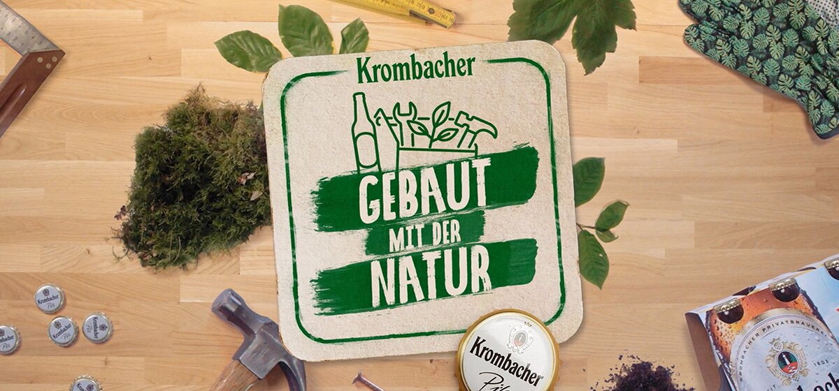 Das Bild zeigt ein Krombacher-Logo mit dem Text 'GEBAUT MIT DER NATUR' auf einem Untergrund aus Holz. Um das Logo sind verschiedene Gartenwerkzeuge, Pflanzen und eine Krombacher-Bierdose angeordnet.