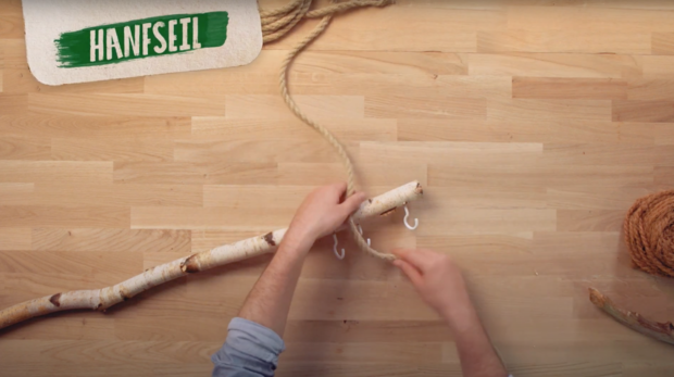 Eine Person bindet ein Hanfseil an einen Holzstock. Auf dem Bild ist ein Teil des Seils und ein Haken sichtbar.