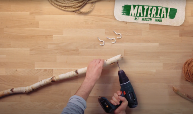 Ein Handwerker hält einen Akkuschrauber in der rechten Hand und bohrt in einen Ast. Auf dem Tisch liegen drei weiße Haken und ein Stück Schnur.