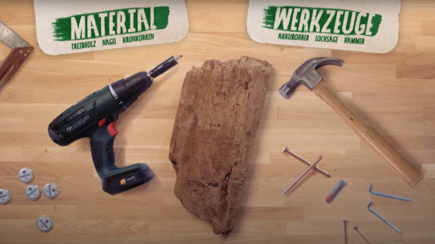 Ein Akkuschrauber, ein Stück Holz, ein Hammer und verschiedene Schrauben sind auf einem Tisch angeordnet. Die Wörter 'MATERIAL' und 'WERKZEUGE' sind in grüner Schrift über den Objekten platziert.
