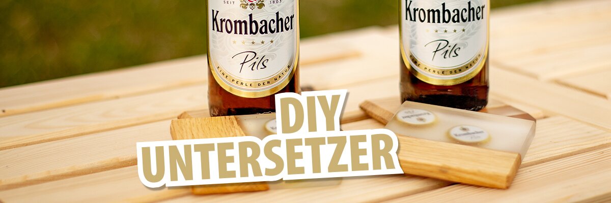 Zwei Flaschen Krombacher Pils stehen auf einem Holztisch. Vor den Flaschen liegen selbstgemachte Untersetzer aus Holz und Kunststoff mit der Aufschrift 'DIY UNTERSETZER'.