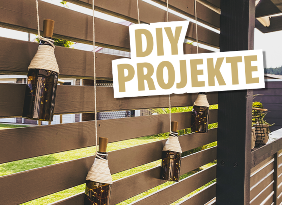 Fünf hängende Flaschen mit Schnur an einer Holzlattenwand. Der Text 'DIY PROJEKTE' ist in großen, hellen Buchstaben über dem Bild platziert.