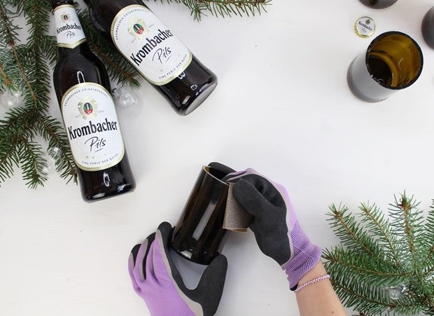 Zwei Hände mit lila Handschuhen halten eine Flasche Krombacher Pils, während sie einen Glasrand mit Schleifpapier bearbeiten. Im Hintergrund sind weitere Krombacher Pils-Flaschen und Tannenzweige zu sehen.