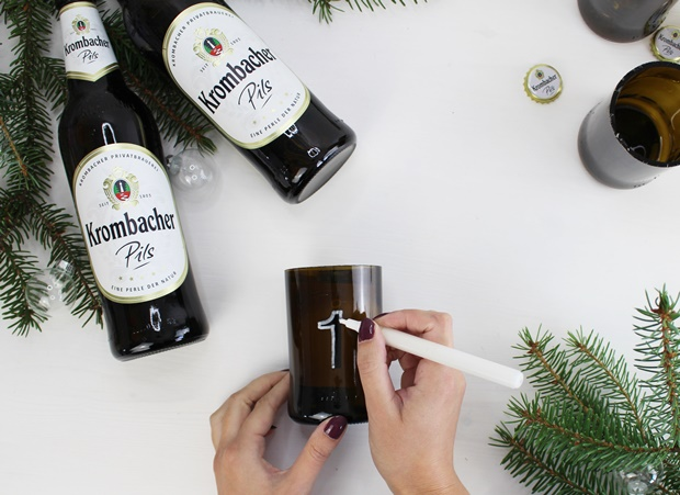 Zwei Flaschen Krombacher Pils stehen neben einem Glas, das mit einer weißen Markierung beschriftet wird. Eine Hand hält einen Stift und zeichnet die Zahl '1' auf das Glas. Im Hintergrund sind Tannenzweige sichtbar.