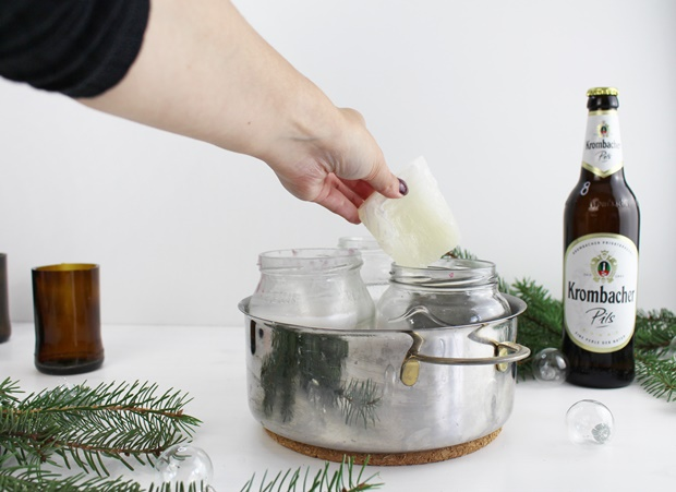 Eine Hand hebt eine weiße Kerze aus einem Glasbehälter in einem silbernen Topf. Daneben steht eine Flasche Krombacher Pils und einige grüne Tannenzweige sind sichtbar.