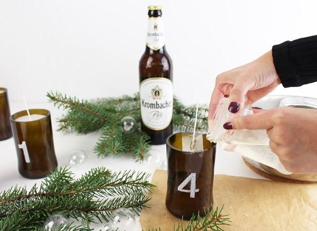 Eine Hand gießt Wachs in ein braunes Glas mit der Nummer '4'. Im Hintergrund steht eine Flasche Krombacher Pils. Um die Gläser liegen Tannenzweige und kleine Dekorationselemente.