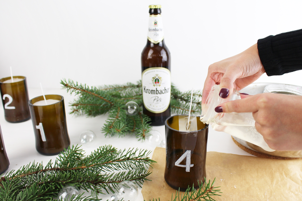 Eine Hand gießt Wachs aus einem Behälter in ein braunes Glas mit der Nummer '4'. Daneben stehen drei weitere braune Gläser mit den Nummern '1', '2' und '3'. Im Hintergrund steht eine Flasche Krumbach Pils und es liegen Tannenzweige sowie transparente Kugeln auf einem braunen Papier.