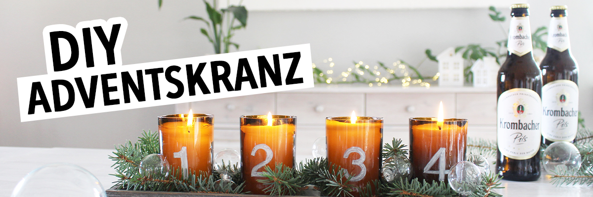Ein Adventskranz aus Holz mit vier brennenden Kerzen in braunen Gläsern, nummeriert von 1 bis 4. Um den Kranz sind Tannenzweige und transparente Kugeln dekoriert. Im Hintergrund stehen zwei Flaschen 'Krombacher Pils'.
