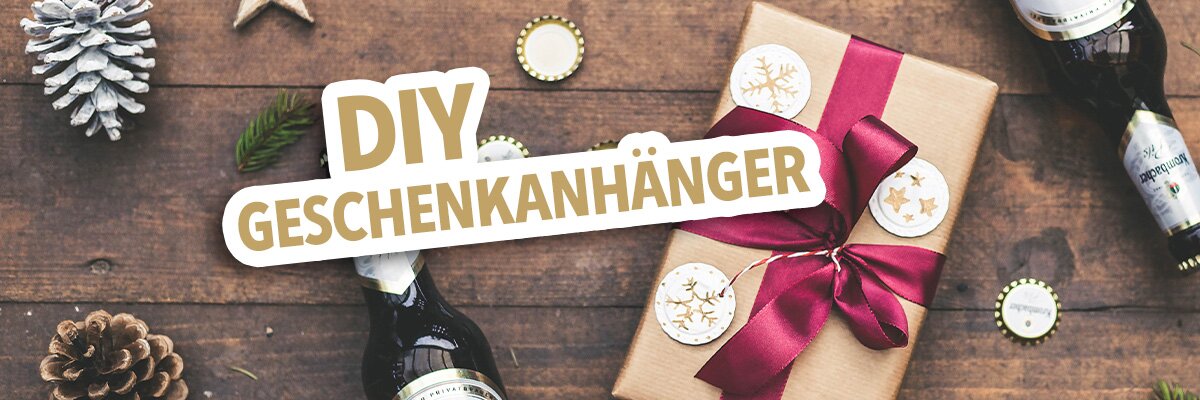 Ein Geschenk mit rotem Band und goldenen Anhängern liegt auf einem Holztisch. Daneben stehen zwei Flaschen Bier und mehrere goldene Sterne.