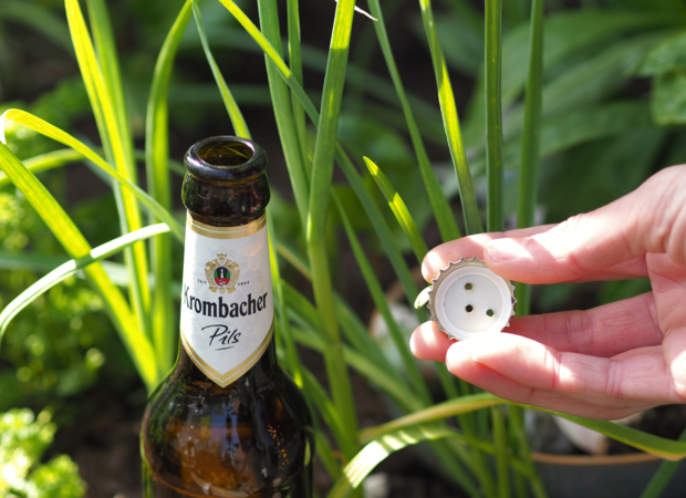 Eine Flasche Krombacher Pils steht zwischen grünen Pflanzen. Eine Hand hält einen Kronkorken mit drei Löchern.
