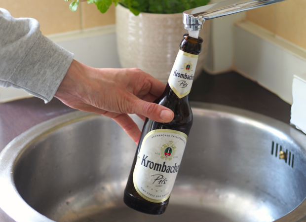 Eine Hand hält eine Flasche Krombacher Pils über einem Spülbecken. Der Wasserhahn ist geöffnet und Wasser fließt in die Flasche.