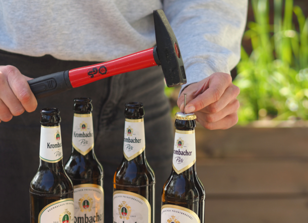 Eine Person hält einen Hammer mit rotem Griff und schlägt auf den Kronkorken einer Krombacher Pils-Flasche. Im Hintergrund sind weitere Krombacher Pils-Flaschen sichtbar.