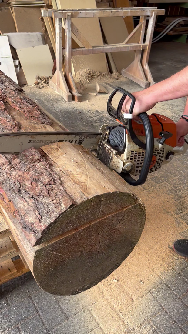 Eine Person hält eine Motorsäge an einem dicken Baumstamm, der auf einem Holzgestell liegt. Der Baumstamm hat eine raue Rinde und ist quer geschnitten.