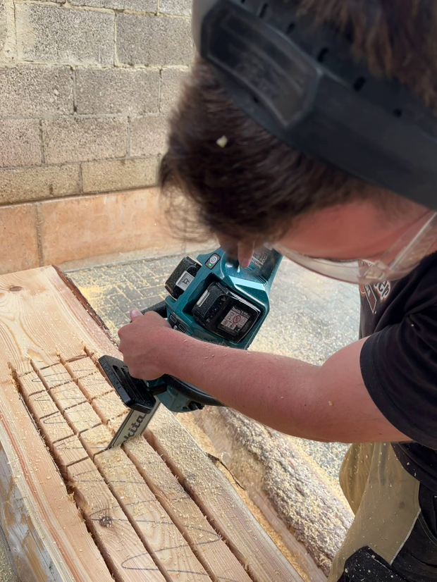Ein Mann mit Schutzbrille und schwarzem T-Shirt schneidet mit einer Makita-Säge in ein Holzbrett. Die Holzoberfläche zeigt Schnittlinien und Späne.