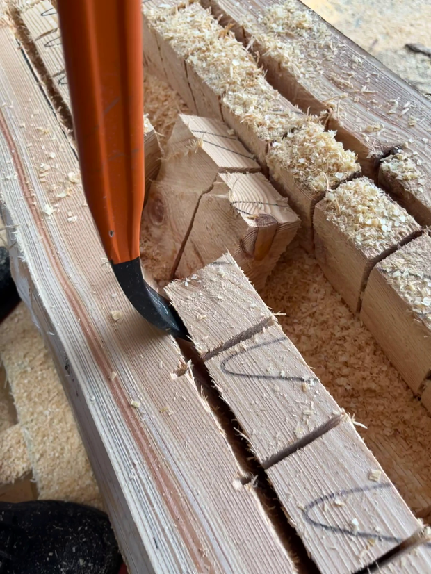 Ein Holzstück mit eingravierten Buchstaben liegt auf einem Tisch, umgeben von Holzspänen. Ein Schnitzwerkzeug mit orangefarbenem Griff ist in der Nähe des Holzstücks platziert.