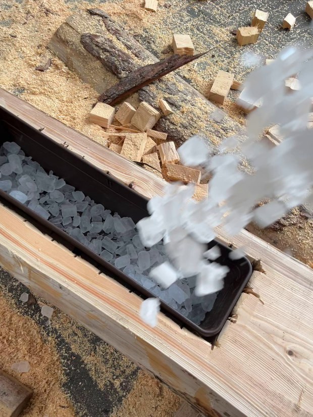 Eiswürfel fallen aus einem Behälter in eine lange, schmale Öffnung aus Holz. Um die Öffnung herum liegen Holzspäne und kleine Holzstücke.