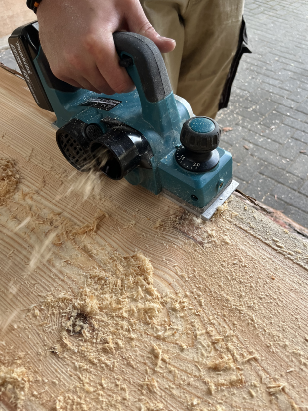 Eine Person benutzt eine elektrische Hobelmaschine in blauer Farbe, um Holz zu bearbeiten. Späne fliegen von der Holzoberfläche, während die Maschine in Bewegung ist.