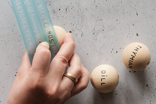 Mit Buchstabenschablone werden Pflanzennamen auf Holzkugeln geschrieben.