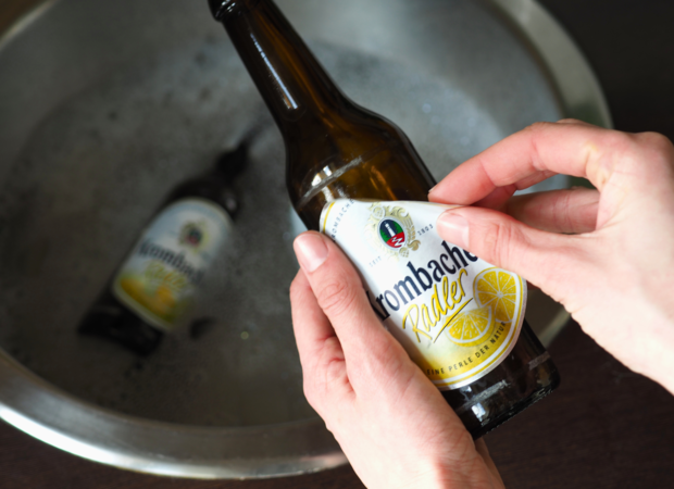 Eine Person hält eine Flasche 'Tombacher Radler' mit gelbem Etikett in der Hand. Im Hintergrund ist eine weitere Flasche im Wasser sichtbar.