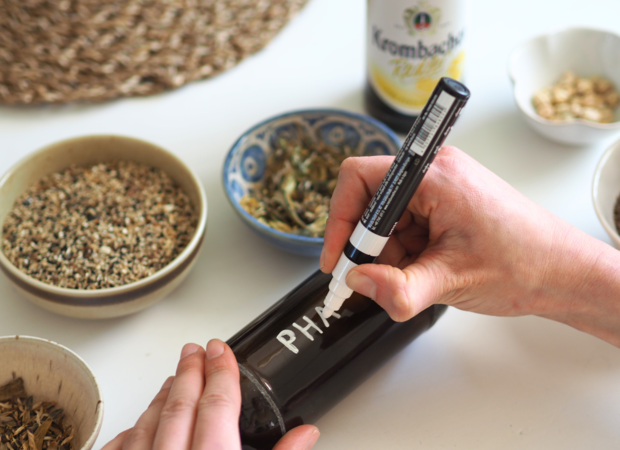 Eine Hand beschriftet eine braune Glasflasche mit dem Wort 'PHARMA' in weißer Farbe. Im Hintergrund sind mehrere Schalen mit verschiedenen Kräutern und eine Flasche Krombacher Pils zu sehen.