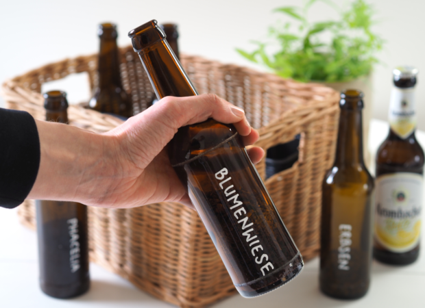 Eine Hand hält eine braune Glasflasche mit dem Etikett 'B.LUMENWIESE'. Im Hintergrund sind weitere Flaschen und ein Korb sichtbar.