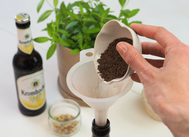 Eine Hand hält eine kleine Schale mit gemahlenem Kaffee über einen Trichter, der in eine Flasche eingesetzt ist. Im Hintergrund steht eine Flasche Krombacher und ein Glas mit Zutaten.