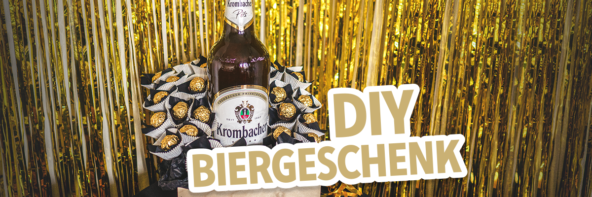 Eine Flasche Krombacher Bier steht in der Mitte eines Geschenks, umgeben von Schokoladenpralinen. Der Text 'DIY BIERGESCHENK' ist in großer, heller Schrift dargestellt.