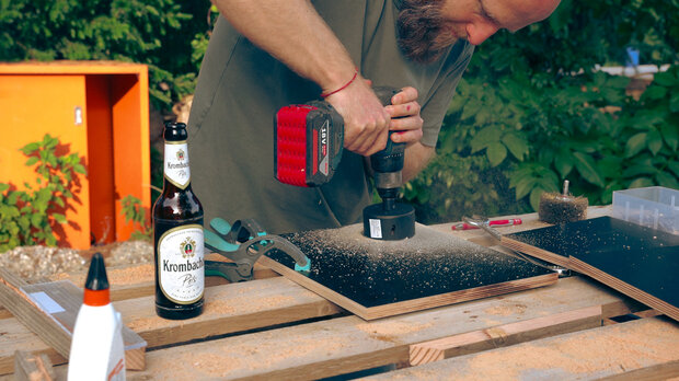 Ein Mann mit Bart bohrt mit einem Akku-Schrauber in eine Holzplatte. Auf einem Holzstapel steht eine Flasche Krombacher Pils. Im Hintergrund sind grüne Pflanzen und ein orangefarbenes Objekt sichtbar.
