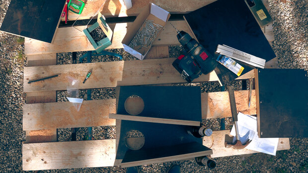 Eine Holzpalette liegt auf einem Kiesboden, darauf sind mehrere schwarze Platten, Schrauben, ein Akkuschrauber, eine Box mit Werkzeugen und einige Papiere verteilt.