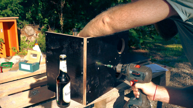 Ein Mann mit einem grauen T-Shirt bohrt mit einem Akku-Schrauber in ein schwarzes Holzgehäuse. Auf einem Holztisch steht eine Flasche Bier und eine grüne Werkzeugkiste.