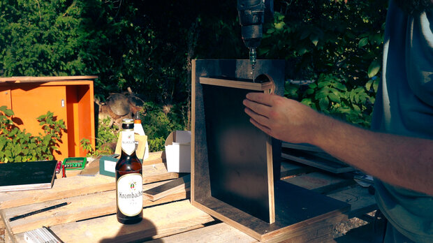 Eine Person mit einem blauen T-Shirt hält ein Holzstück an einem schwarzen Kasten, während sie mit einem Bohrer arbeitet. Auf einem Tisch steht eine Flasche Kombucha-Bier.
