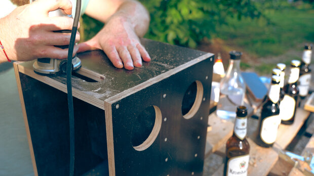 Ein Handwerker bearbeitet einen Holzblock mit einem Schleifgerät. Der Block hat zwei runde Aussparungen und steht auf einer Holzpalette. Im Hintergrund sind mehrere Flaschen, darunter 'Kunstmann', sichtbar.
