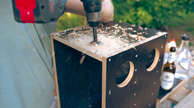 Ein Handwerker bohrt mit einem Akkuschrauber ein Loch in eine schwarze Holzkiste mit zwei runden Öffnungen. Späne fliegen auf die Oberfläche. Im Hintergrund sind Flaschen sichtbar.