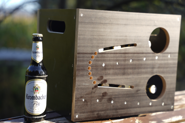 Eine Kiste aus Holz mit zwei runden Ausschnitten und mehreren kleinen Löchern steht neben einer Flasche Krombacher Pils. Die Kiste hat eine rechteckige Form und eine glatte Oberfläche.